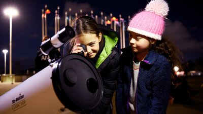 1 child stares through a telescope outside Parliament House while another child stands beside them.