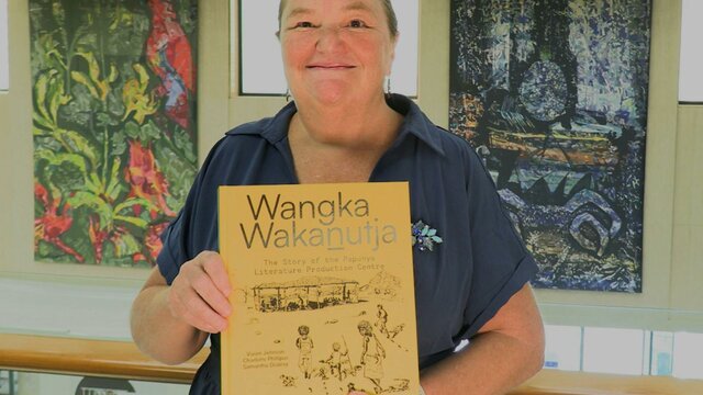 A woman holding a book with a yellow cover, standing in front of some wall tapestries