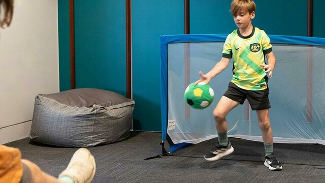 Little Kickers at the National Portrait Gallery A child kicks a soccer ball towards a goal, where a child in a Matildas Jersey defends the ball.