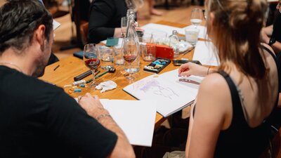 People sitting at a table drawing, with glasses of red wine