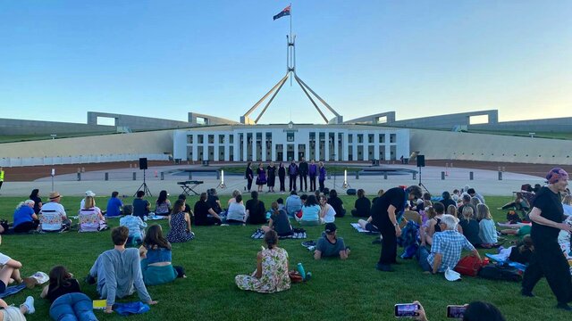 A group of people sitting on the lawns of Australian Parliament House watching a choir.