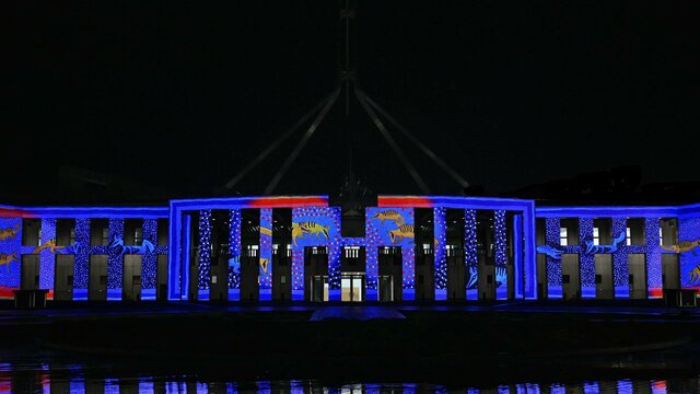 Parliament House at night, illuminated in blue & red light with projected artwork across the façade