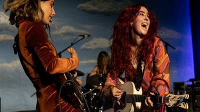 2 young woman dressed in fun outfits play guitars and sing against a sky blue backdrop