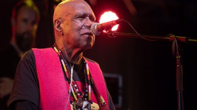 Uncle Archie roach sings on stage, wearing a black shirt and red waistcoat