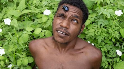 A photographic portrait of a First Nations man siting amongst foliage with a beetle on his forehead.