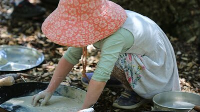 A young child wearing a sunhat, mixing dirt and water in an old frying pan