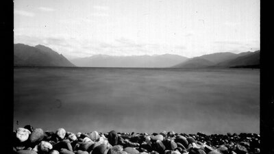 A black and white photograph of a pebbly beach and mountains in the background