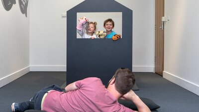 Two children playing in a puppet theatre while man watched on cushions