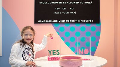 Girl putting a pink token in a bucket to vote on a question on the wall
