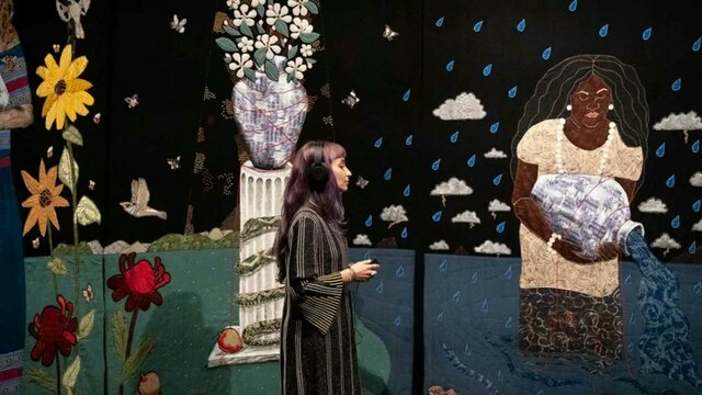 Tal Fitzpatrick | Quietly Seething: Fate, Fury and Transmutation A woman stands in front of a large tapestry artworks with flowers and a woman with a water vessel