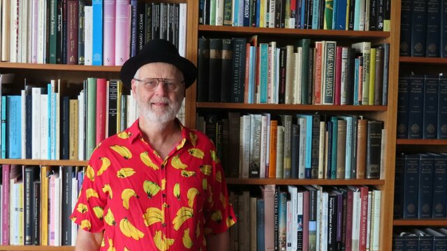 Prof Matthew Spriggs An elderly man wearing a black hat and a banana printed shirt while standing in front of a bookcase
