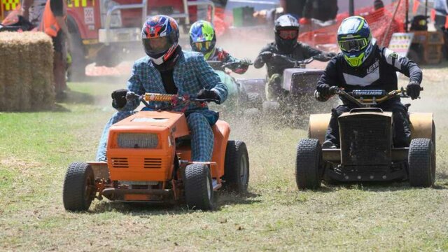 nla.obj-3640189333 Competitors race with lawnmowers on a grass track with a fire truck and spectators visible.