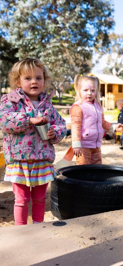 Young children play in a sandpit and look at the camera.