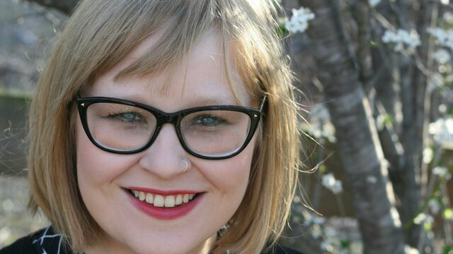 Dr Meg Brayshaw A smiling woman with glasses and blonde hair stands outdoors near blooming trees.