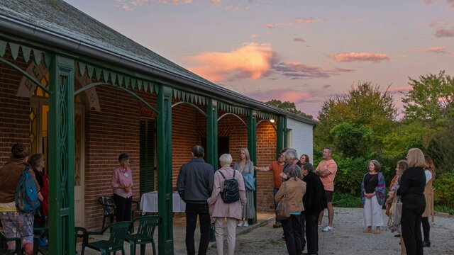 People gather on the verandah of a historic red brick house. The sun sets above.