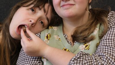 Two young women sit next to each other. One of the women feeds the other a chocolate.