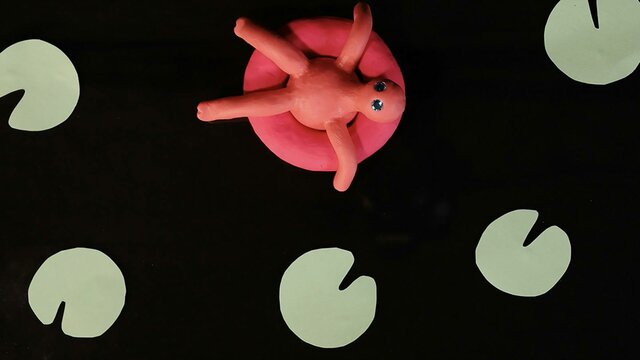 A video still image of a pink clay character sitting in a pool float surrounded by green lily-ads