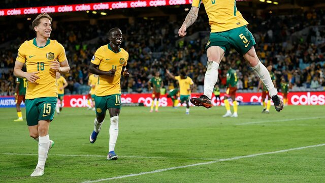 Soccer players in yellow kits celebrate a goal on a stadium pitch as teammates run in behind