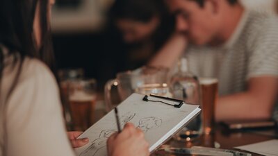 Person drawing in foreground, with glasses of beer on table