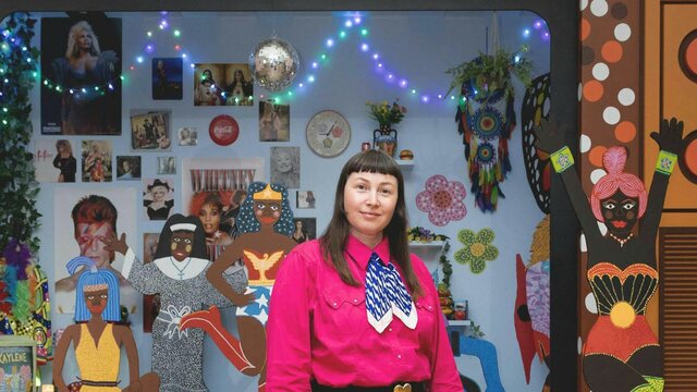 April Phillips with Kaylene TV, 2023. © Kaylene Whiskey A person wearing a pink shirt stands in front of an 3D artwork featuring a disco ball and figures.