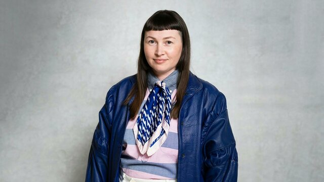 April Phillips A person wearing a scarf, striped shirt, and blue leather jacket stands against a plain grey wall.