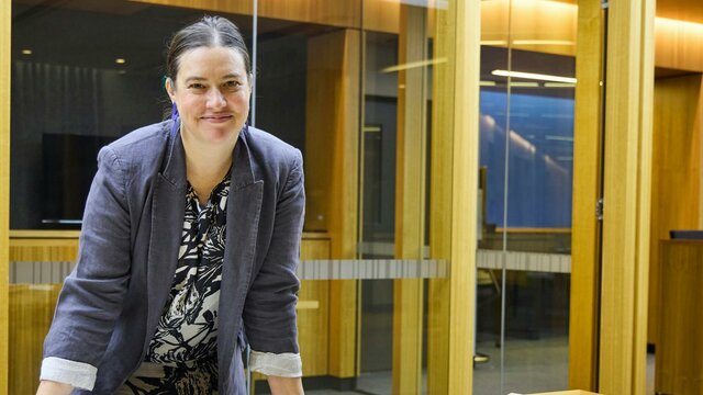 Catherine Dwyer A woman in a grey blazer smiles at the camera while leaning on a wooden table covered with books.