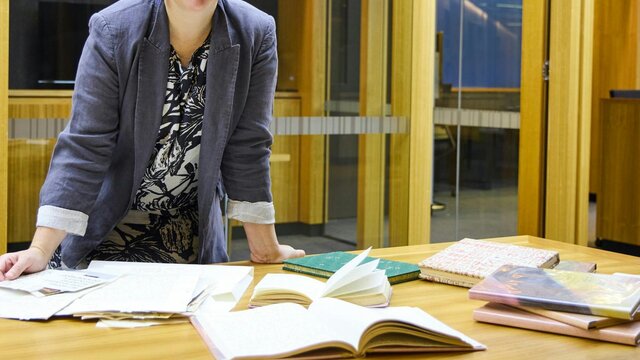 Catherine Dwyer A woman in a grey blazer smiles at the camera while leaning on a wooden table covered with books.