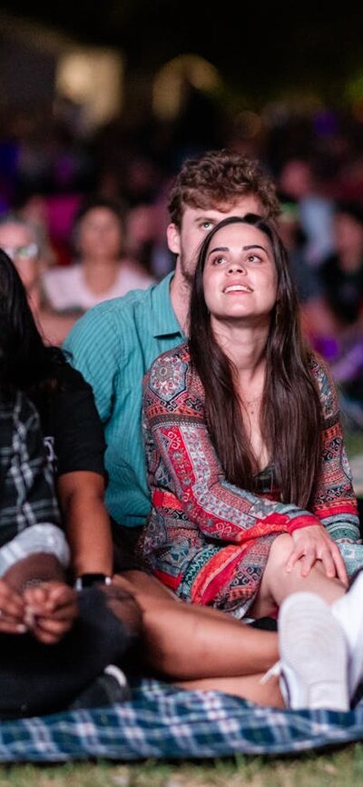 People sitting on grass, looking up at stage as they watch outdoor performance