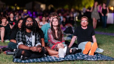 People sitting on grass, looking up at stage as they watch outdoor performance