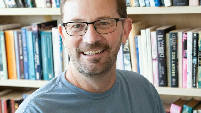 Anthony Eaton Man with glasses smiles in front of bookshelf