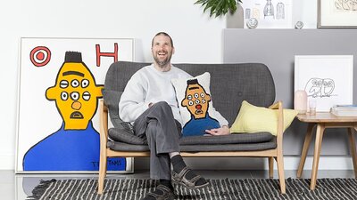 Thom Roberts with Burt the Oscar Train 2017 and cushion cover. A man sitting on grey couch holding a colourful cushion.