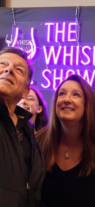 A group gathers in front of a Whisky Show banner and neon sign for a selfie.