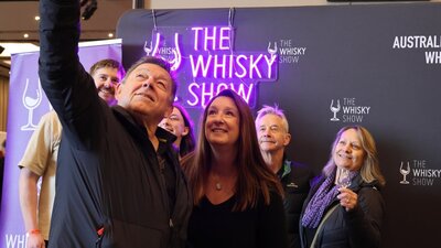 A group gathers in front of a Whisky Show banner and neon sign for a selfie.