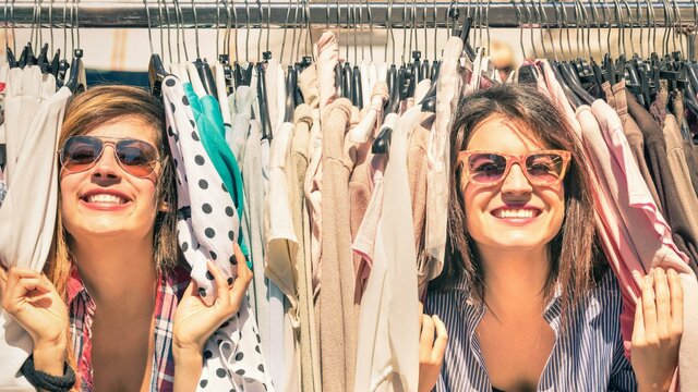 two women smiling peeking through a rack of clothes at an outdoor market