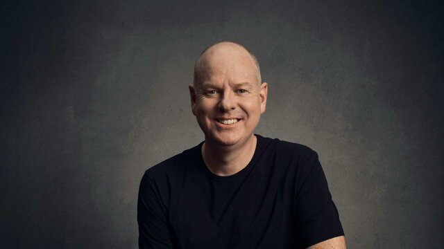 Comedian Tom Gleeson sitting on a stool, smiling