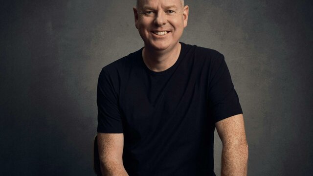 Comedian Tom Gleeson sitting on a stool, smiling