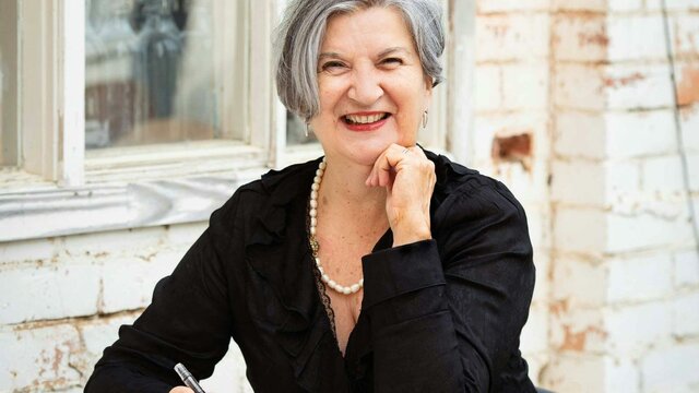 Bobby Graham A smiling woman with grey hair sits at a table outdoors, writing in a notebook.
