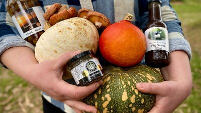 A person holds colourful pumpkins, jar of olives, black garlic, fig sauce and mushrooms.