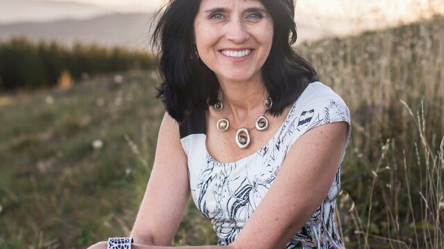 Karen Viggers A woman with dark, wavy hair sitting outdoors, wearing a white patterned top.