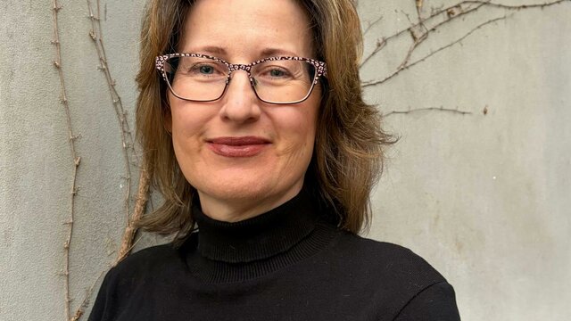 Claudia Craig A woman with brown, wavy hair and glasses, wearing a black turtleneck.