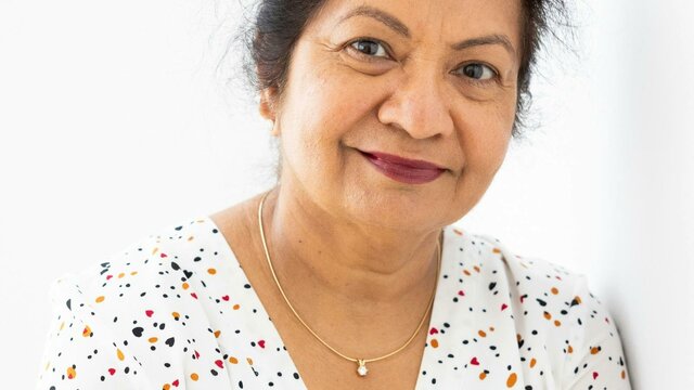 Neera Mahajan A woman smiling while resting her right hand on her left shoulder, while wearing a polka dot shirt.