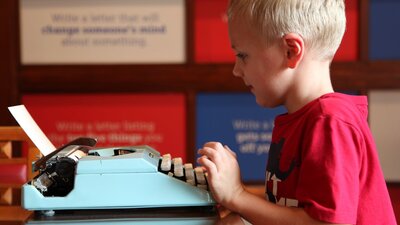 Young boy in red shirt typing at light blue typewriter in MoAD’s Yours Faithfully exhibition space