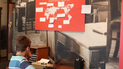 Boy thinking at typewriter in front of red and white world map showing visitor’s travel stories