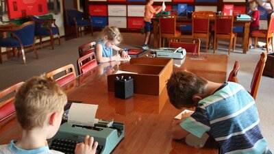 Three children at a wooden table writing letters by hand and on a typewriter