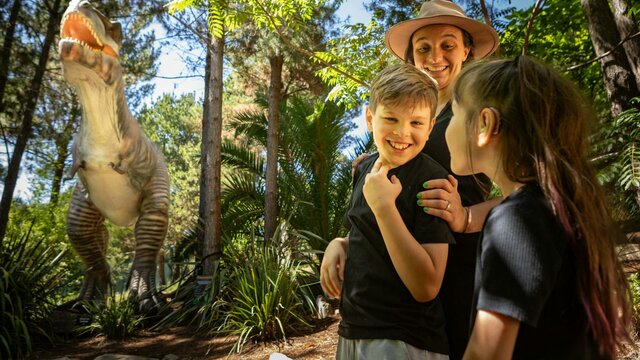 Extinction Trail - TREX A mother and two children smile in awe at the size of the animatronic T-Rex in lush fern gardens.