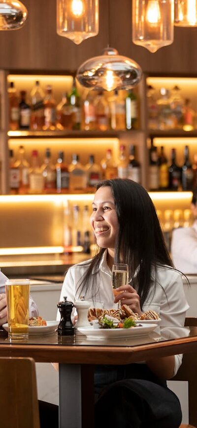 Two tables of guests enjoying drinks while a smiling bartender in the background shakes a cocktail