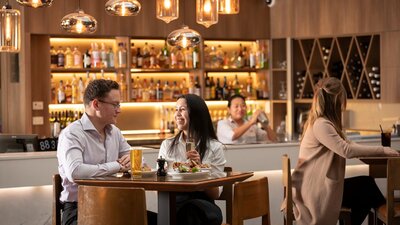 Two tables of guests enjoying drinks while a smiling bartender in the background shakes a cocktail