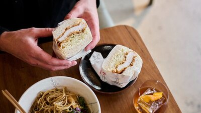 Hands holding a fluffy katsu sandwich with a bowl or ramen and a drink nearby.