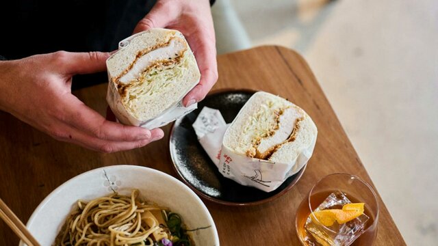 Hands holding a fluffy katsu sandwich with a bowl or ramen and a drink nearby.