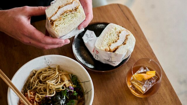 Hands holding a fluffy katsu sandwich with a bowl or ramen and a drink nearby.